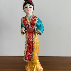 Op de afbeelding: Een decoratieve pop met zwart haar in een knotje met een gouden en roze bloemornament. De pop draagt een kleurrijke outfit met een rood patroon op het lijfje, blauwgroene mouwen, een bruine tailleband en een lange gele rok met roze bloemenprints.
