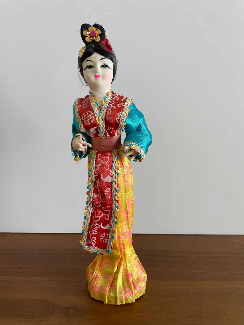 Op de afbeelding: Een decoratieve pop met zwart haar in een knotje met een gouden en roze bloemornament. De pop draagt een kleurrijke outfit met een rood patroon op het lijfje, blauwgroene mouwen, een bruine tailleband en een lange gele rok met roze bloemenprints.