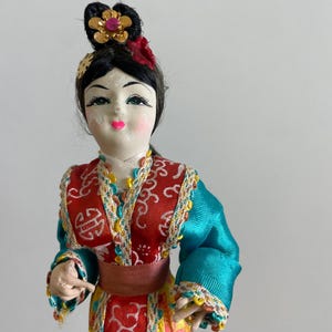 Op de afbeelding: Een decoratieve pop met zwart haar in een knotje met een gouden bloemornament. De pop draagt een kleurrijke outfit met een rood lijfje, blauwgroene mouwen en een gele rok, afgewerkt met ingewikkelde witte patronen en kleurrijke randen.