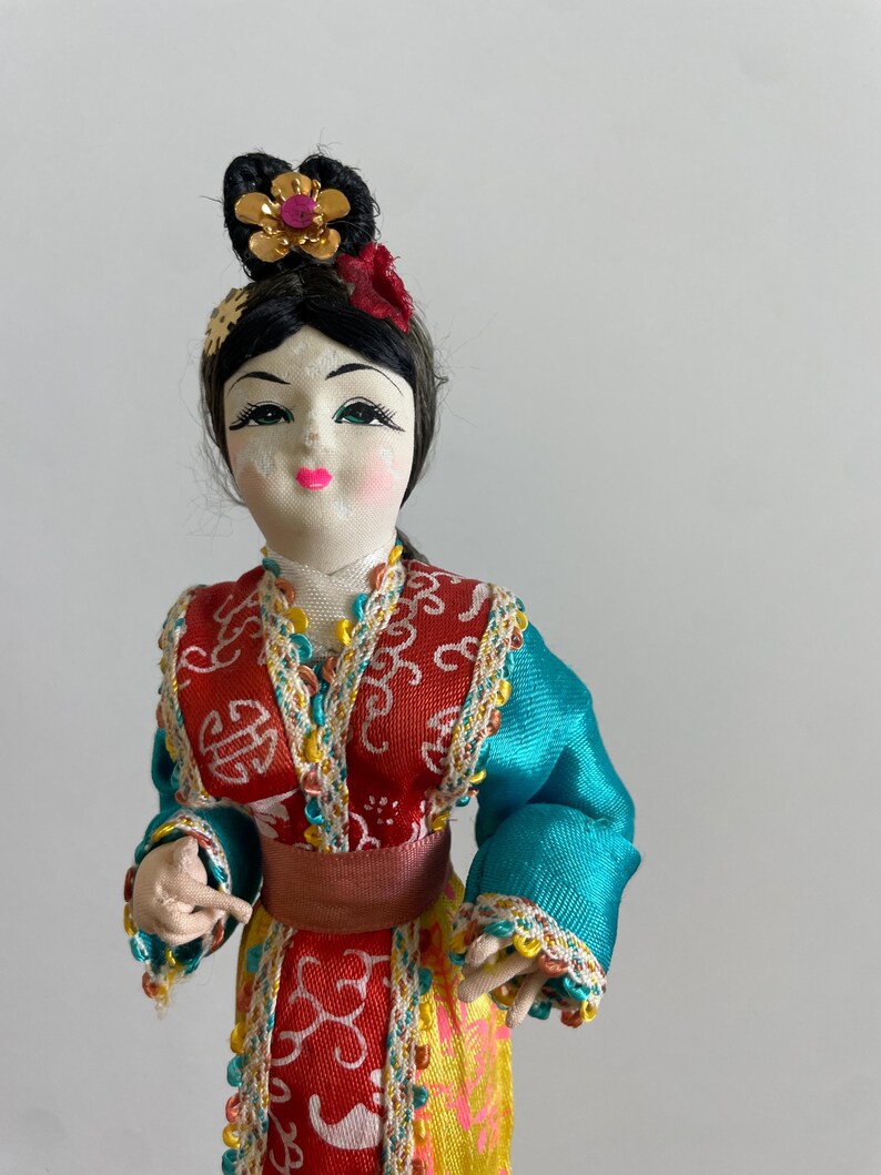Op de afbeelding: Een decoratieve pop met zwart haar in een knotje met een gouden bloemornament. De pop draagt een kleurrijke outfit met een rood lijfje, blauwgroene mouwen en een gele rok, afgewerkt met ingewikkelde witte patronen en kleurrijke randen.
