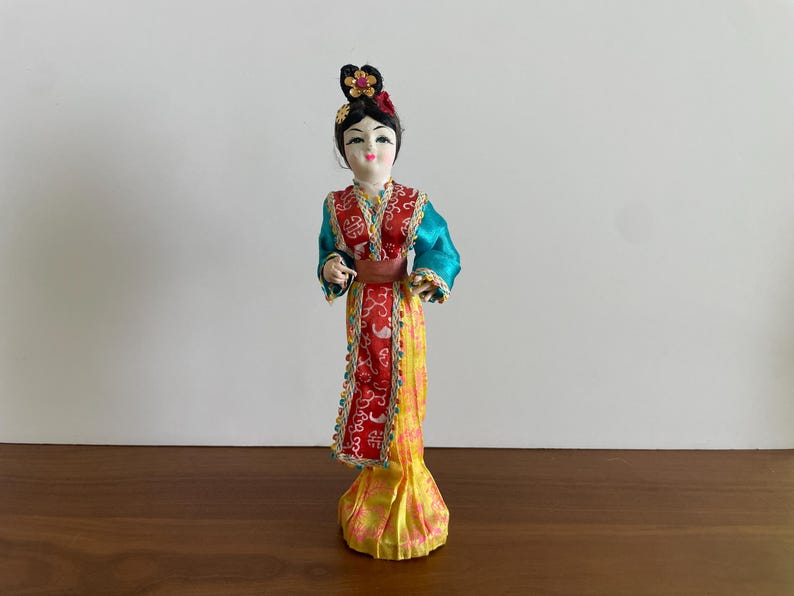 Op de afbeelding: Een decoratieve pop met donker haar in een opgestoken kapsel met een gouden en roze bloemenornament. De pop draagt een lange jurk met een blauwgroen lijfje, een rood middenpaneel met witte patronen en een gele rok met een roze bloemenprint.