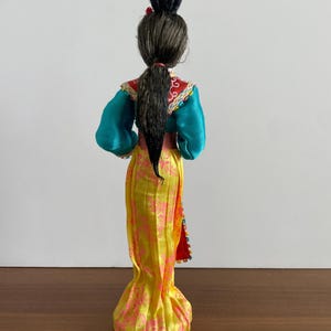 Op de afbeelding: Een pop met donker haar in een hoge knot en een paardenstaart, gekleed in een groenblauw shirt met lange mouwen en een rode kraag, en een lange gele rok met een roze bloemenpatroon. De outfit is afgewerkt met decoratieve randen.