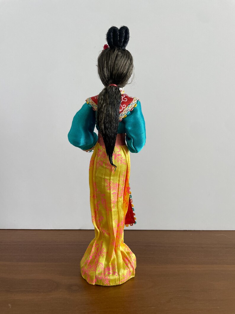 Op de afbeelding: Een pop met donker haar in een hoge knot en een paardenstaart, gekleed in een groenblauw shirt met lange mouwen en een rode kraag, en een lange gele rok met een roze bloemenpatroon. De outfit is afgewerkt met decoratieve randen.