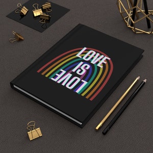 Peut inclure: Un carnet noir avec un motif arc-en-ciel et le texte "LOVE IS LOVE" sur la couverture. Le carnet est posé sur une surface grise avec des trombones dorés et des crayons.