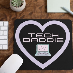 Może przedstawiać: Czarna podkładka pod mysz w kształcie fioletowego serca, zawierająca laptop i tekst "TECH BADDIE".