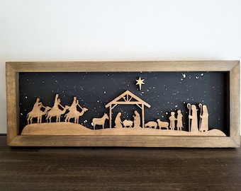May include: A wooden framed shadow box with a black background and a gold star. The shadow box depicts the nativity scene with three wise men riding camels, a manger with a baby Jesus, and shepherds.