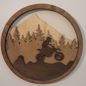 May include: A round wooden wall art piece featuring a motocross rider on a dirt bike in a mountain landscape. The design is layered with a dark brown frame, a light brown mountain and trees, and a darker brown foreground.
