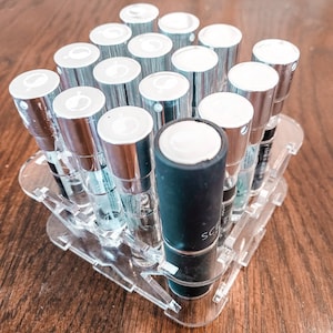 May include: A clear acrylic display stand with 15 small glass bottles, each with a silver cap. The bottles are arranged in a grid pattern and contain a clear liquid. One bottle is a dark grey colour with a black cap.