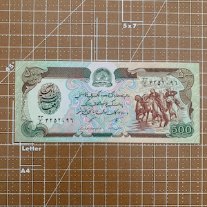 Può includere: Una banconota afghana di 500 afghani, di colore verde e marrone. La banconota presenta una scena di persone a cavallo e un sigillo con le parole "Da Afghanistan Bank" in inglese e dari.