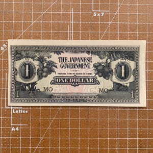 May include: A vintage Japanese Government one-dollar bill. The bill features the text "THE JAPANESE GOVERNMENT" and "ONE DOLLAR". The design includes illustrations of fruit and the number "1" in two ornate circles.