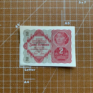May include: A vintage Austrian 2 Kronen banknote. The note features a portrait of a woman and ornate red and gray designs. The text "Zwei Kronen" is visible, along with other German text.