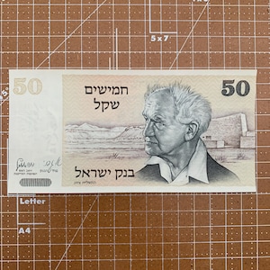 May include: A fifty Israeli shekel banknote. The bill features a portrait of a man on the right side, with Hebrew text and the number 50 on both sides. The background shows a landscape.