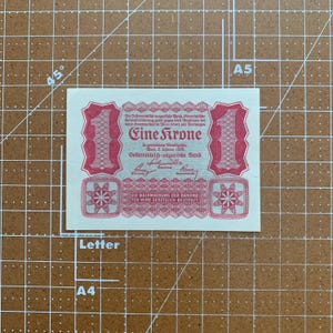 May include: A vintage Austrian banknote, featuring the text "Eine Krone" in red ink. The design includes ornate borders, a large "1" on each side, and text in German. The note is on a grid background.
