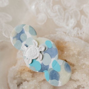 May include: A decorative hair clip featuring a white turtle on a blue, white, and light blue polka-dot design. The clip is made of resin and is approximately 3 inches long.