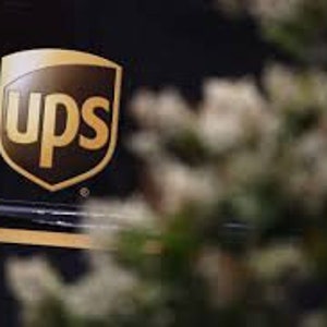 May include: The UPS logo is a brown shield with the letters "UPS" in white, surrounded by a gold border. The logo is on a black background.