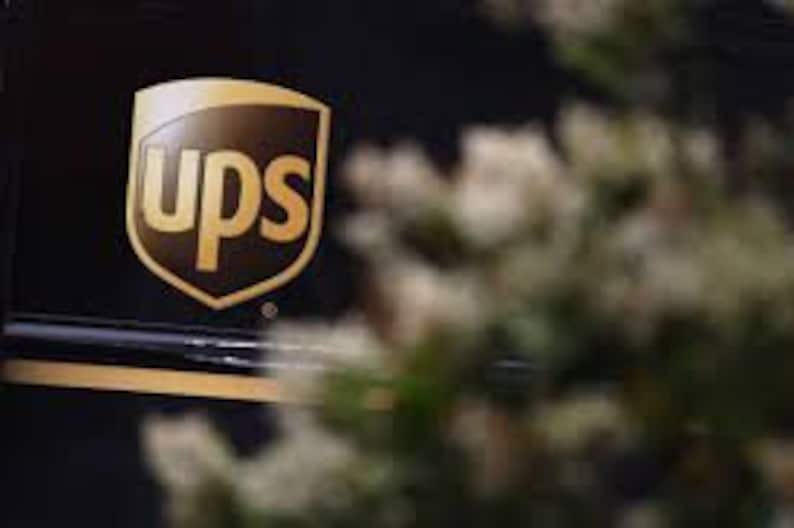May include: The UPS logo is a brown shield with the letters "UPS" in white, surrounded by a gold border. The logo is on a black background.