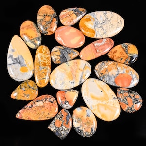 以下が含まれることがあります： 様々な形に研磨された宝石のコレクション。楕円形や涙型など。オレンジ、ピーチ、グレー、黒の色合いが混ざり合い、ユニークな模様と質感があります。宝石は黒い表面に配置されています。