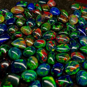 May include: A collection of iridescent black opal gemstones, each with a unique pattern of green, blue, and red colours. The opals are cut in a teardrop shape and have a smooth, polished finish.