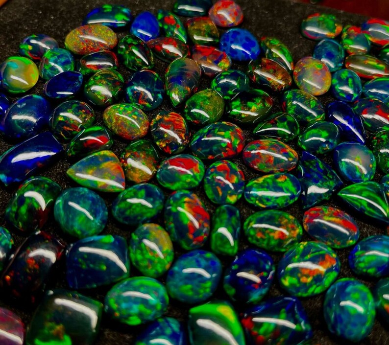 May include: A collection of iridescent black opal gemstones, each with a unique pattern of green, blue, and red colours. The opals are cut in a teardrop shape and have a smooth, polished finish.