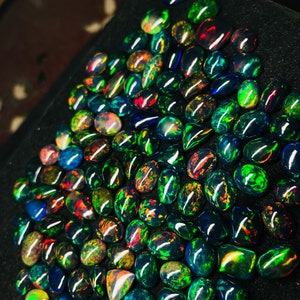 May include: A large collection of black opal gemstones, with iridescent green, blue, and red hues. The opals are displayed on a black surface.