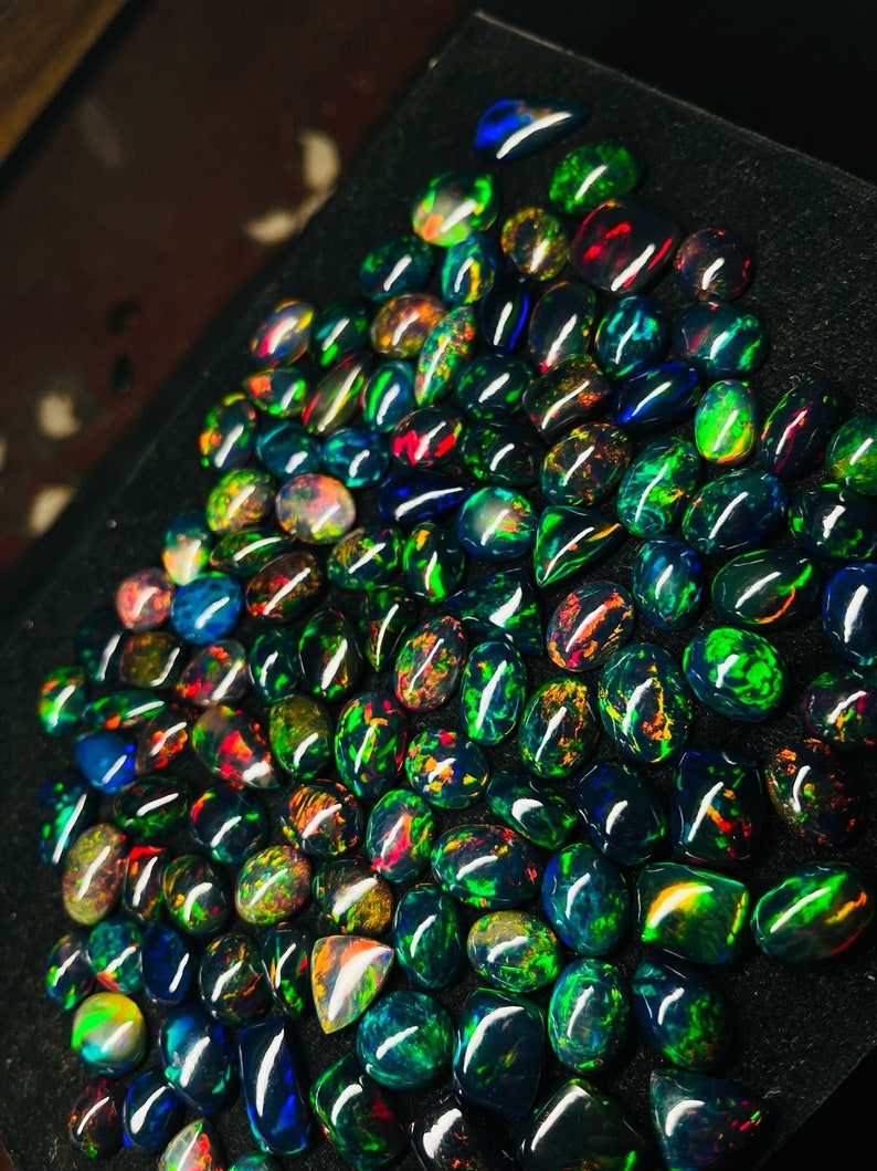 May include: A large collection of black opal gemstones, with iridescent green, blue, and red hues. The opals are displayed on a black surface.