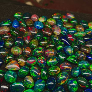 May include: A collection of black opal gemstones with iridescent green, blue, and red hues. The opals are displayed on a black surface.