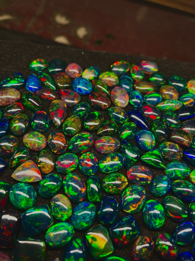 May include: A collection of black opal gemstones with iridescent green, blue, and red hues. The opals are displayed on a black surface.
