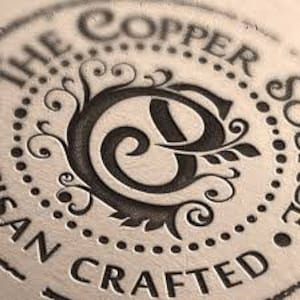 May include: A close-up of a circular, copper-coloured stamp with the text "The Copper Spoon" and "Hand Crafted" around the edge. The centre of the stamp features a stylised letter "C" with a floral design.