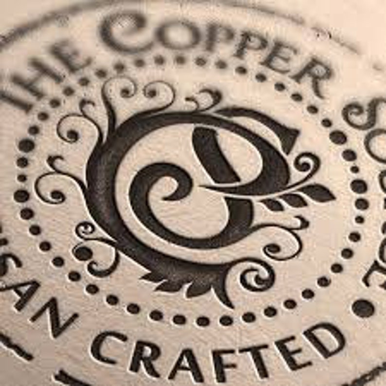 May include: A close-up of a circular, copper-coloured stamp with the text "The Copper Spoon" and "Hand Crafted" around the edge. The centre of the stamp features a stylised letter "C" with a floral design.
