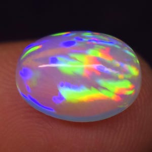 May include: An oval-shaped cabochon opal gemstone featuring a translucent white base with vibrant flashes of neon green, yellow, orange, and blue color play across its smooth, polished, and domed surface against a dark background.