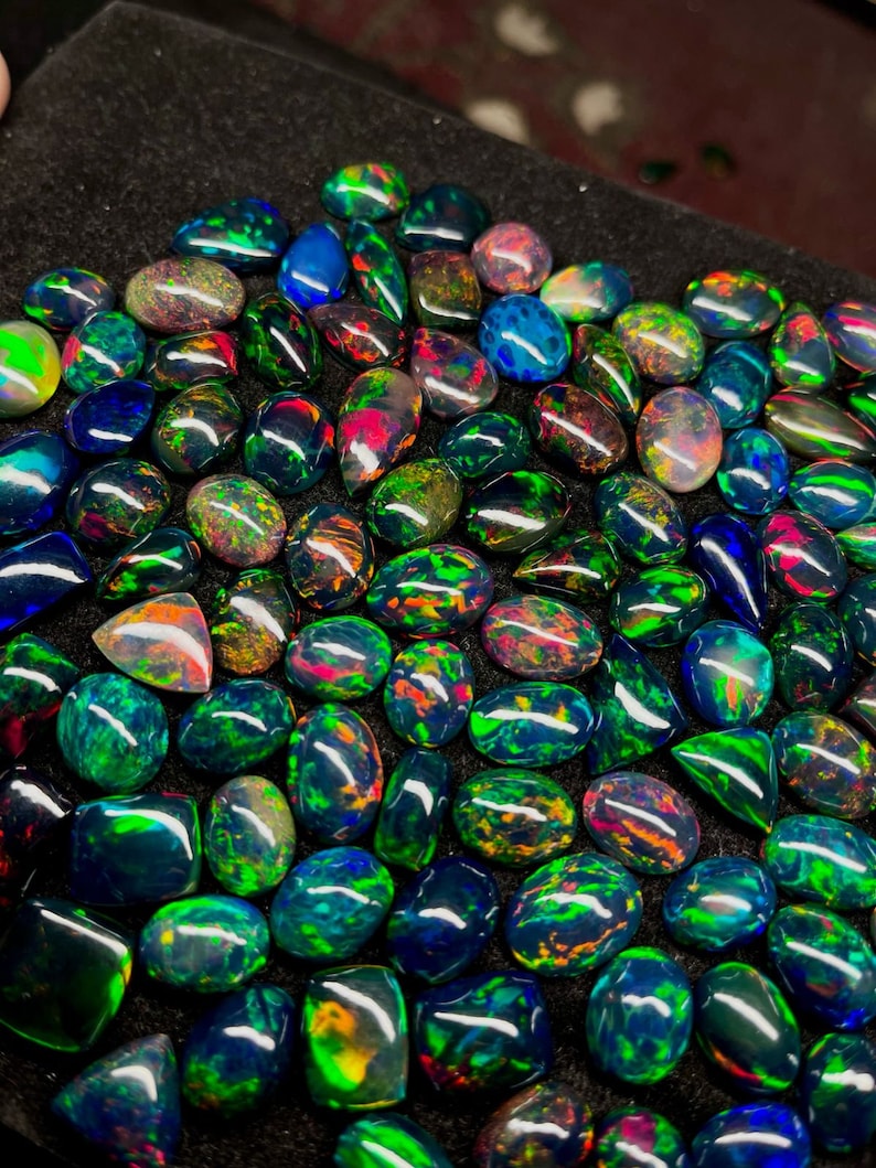May include: A collection of black opal gemstones with iridescent green, blue, and red colours. The opals are displayed on a black surface.