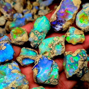 Può includere: Una collezione di opali grezzi, che mostrano colori vivaci come blu, verde e arancione. Gli opali sono tagliati in modo grezzo, con una matrice naturale e terrosa. Le pietre preziose sono tenute in una mano, evidenziando le loro dimensioni e brillantezza.