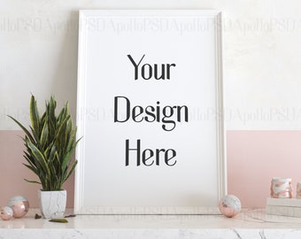 May include: A white framed print with the text "Your Design Here" is displayed on a marble surface. A potted snake plant, decorative spheres, and small decorative jars are also on the surface. The background has a pink and white color scheme.