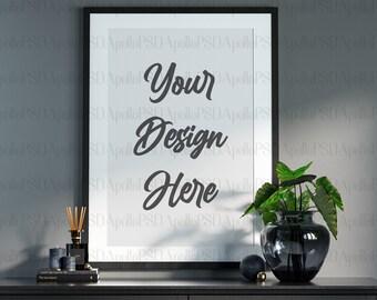May include: A black framed poster with the text "Your Design Here" on a white background. The poster is on a black table with a vase of green plants and a book.