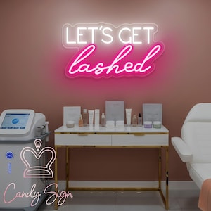Puede incluir: Interior de un salón de belleza con una pared rosa y un letrero de neón que dice "LET'S GET lashed". Una mesa blanca y dorada exhibe productos de belleza. También se ven una silla de tratamiento blanca y un dispositivo médico.