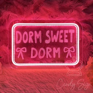 Może przedstawiać: Różowy neon z napisem "DORM SWEET DORM" w prostokątnej ramie. Znak zawiera grafiki kokardek i jest umieszczony na różowym, pierzastym tle. To element dekoracyjny.