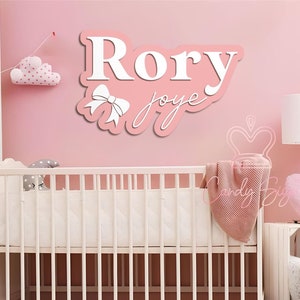 May include: Pink and white wooden name sign with the name "Rory" and the word "joye" written in cursive. The sign is hanging on a pink wall above a white cot.