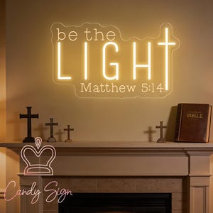 May include: A wall-mounted neon sign featuring the text "be the LIGHT" in a warm yellow glow, with the letter T replaced by a cross. Below the text, the words "Matthew 5:14" are displayed in a thinner, white font style on the sign.