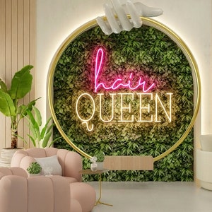 May include: A pink neon sign reading "hair queen" is mounted on a wall covered in green plants. The sign is surrounded by a gold circle. There are two pink chairs in the foreground.