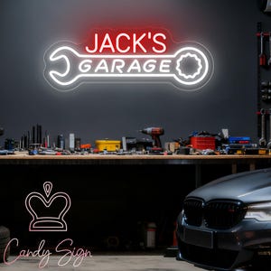 Può includere: Un'insegna al neon per "Jack's Garage" con una combinazione di colori rosso e bianco. L'insegna ha la forma di una chiave inglese ed è montata su una parete grigio scuro. L'ambiente del garage include attrezzi, un banco da lavoro e un'auto.