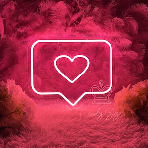 May include: Pink neon sign shaped like a speech bubble with a white heart inside. The sign is on a pink fluffy surface with pink feathers in the background.