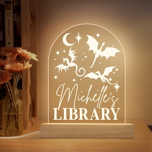 May include: A glowing acrylic night light with a wooden base. The light features a dragon and star design with the words "Michelle's LIBRARY" in a decorative font. The light is on a wooden table with a vase of flowers in the background.