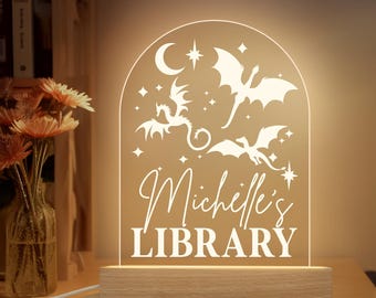 Op de afbeelding: Verlicht acryl nachtlampje met een houten voet. Het ontwerp toont draken en sterren, met de woorden "Michelle's LIBRARY". Het licht staat op een houten tafel. Een vaas met bloemen is zichtbaar op de achtergrond.