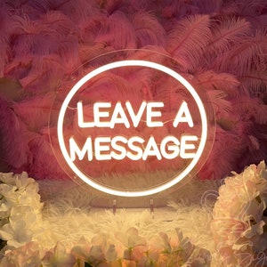 Op de afbeelding: Een wit neonbord met een heldere ronde lijst, waarop de tekst "LEAVE A MESSAGE" staat tegen een roze veren achtergrond.