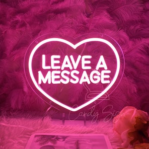 Puede incluir: Un letrero de neón rosa en forma de corazón con las palabras "LEAVE A MESSAGE" en letras de neón blancas.