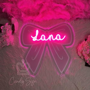 May include: A pink neon sign in the shape of a bow with the name "Lana" in cursive script. The sign is surrounded by a fluffy pink background and the words "Candy Sign" are at the bottom.