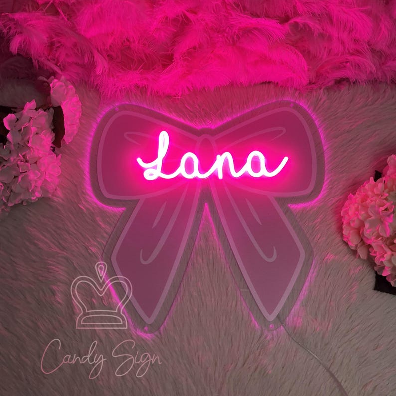 May include: A pink neon sign in the shape of a bow with the name "Lana" in cursive script. The sign is surrounded by a fluffy pink background and the words "Candy Sign" are at the bottom.