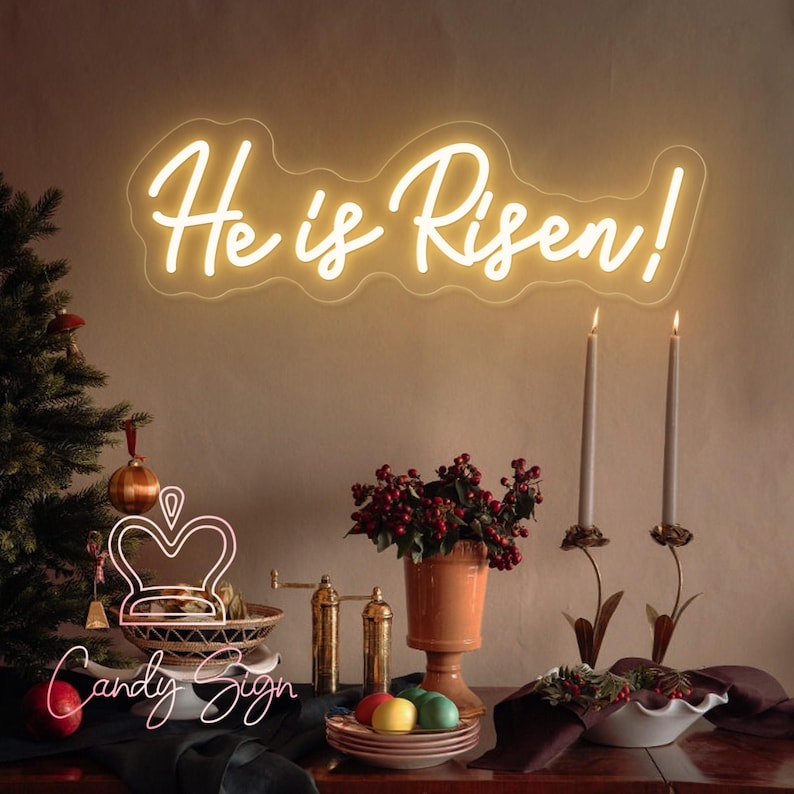 May include: White neon sign with the text "He is Risen!" on a beige wall. The sign is surrounded by a decorative arrangement of candles, a vase with red berries, and a bowl of Easter eggs.