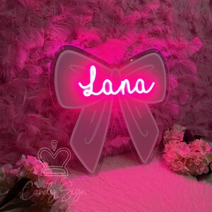 May include: A pink neon sign in the shape of a bow with the name "Lana" in a cursive font. The sign is mounted on a pink feathery background. The sign is from "Candy Sign".