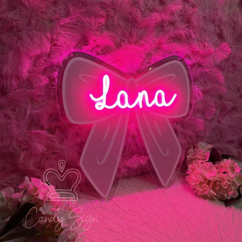 May include: A pink neon sign in the shape of a bow with the name "Lana" in a cursive font. The sign is mounted on a pink feathery background. The sign is from "Candy Sign".